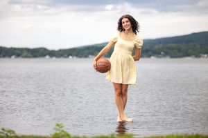 Young woman in yellow dress holding basketball standing barefoot in shallow lake water with scenic hills in background.