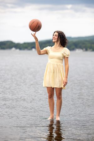 Young woman in yellow dress spinning basketball on finger while standing in lake water.