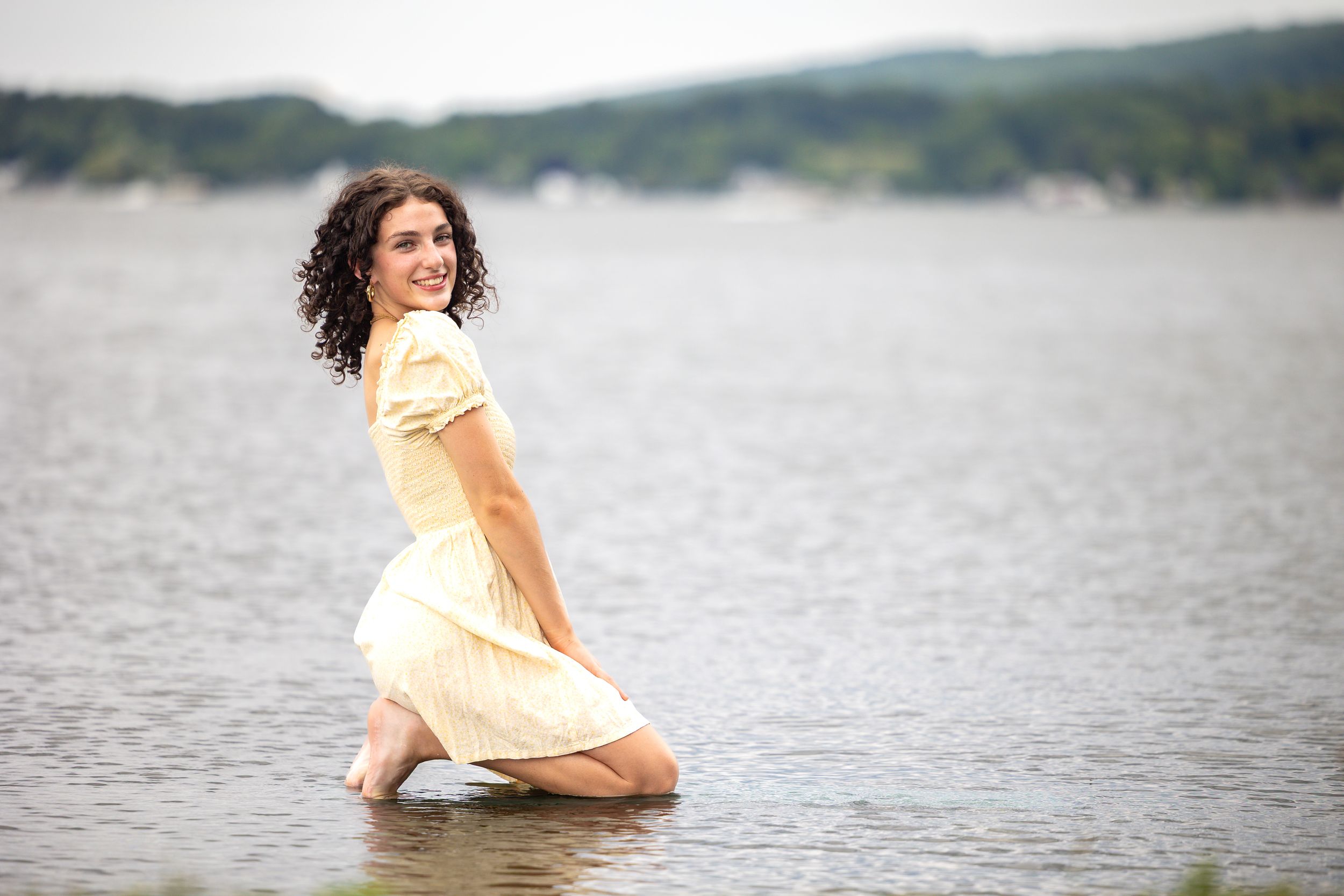 Young woman in yellow dress kneeling in lake water smiling.