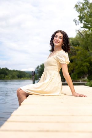 Smiling young woman in yellow dress sitting on wooden dock by lake.