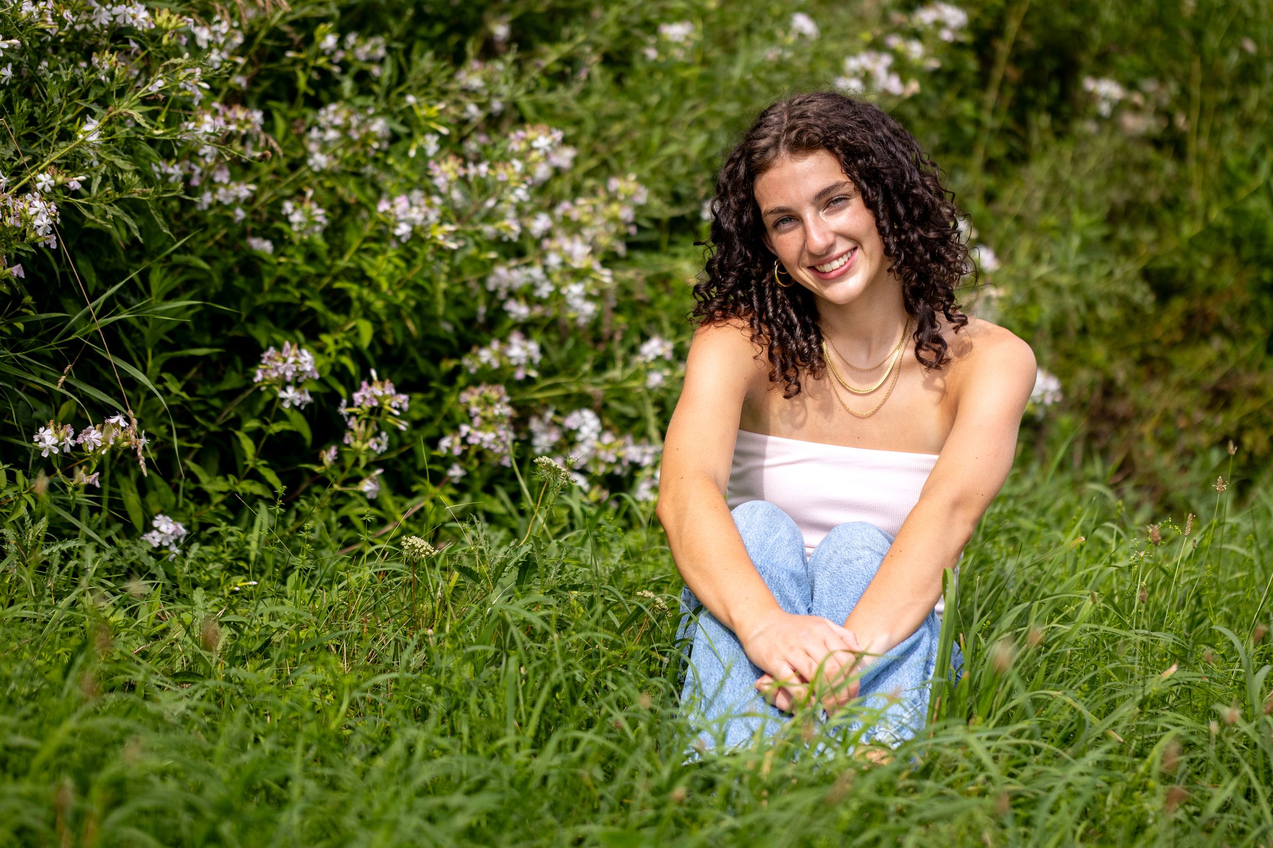 Young woman in white top and blue skirt sitting in green grass with wildflowers.