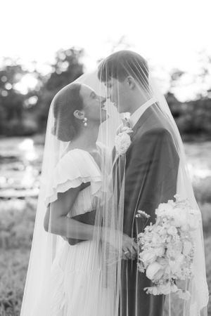 A romantic black and white wedding photo of a couple in formal attire with a flowing veil and bouquet of flowers outdoors.