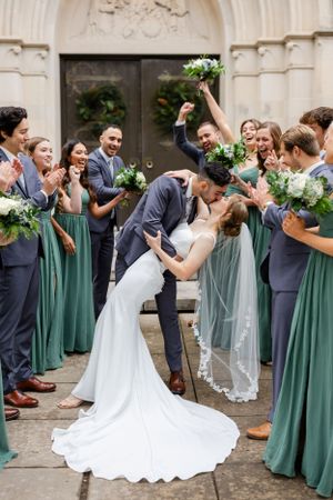 Wedding couple shares a romantic dip kiss surrounded by bridesmaids in sage green dresses and groomsmen in navy suits.