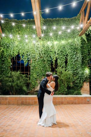 String lights illuminate wedding dance sequence on outdoor terrace surrounded by lush greenery and brick walls.