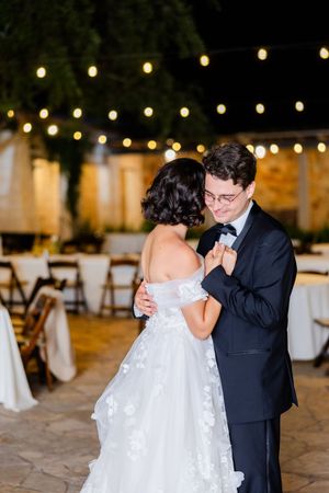 A couple shares their first dance on a stone patio with romantic string lights glowing overhead at an outdoor reception.