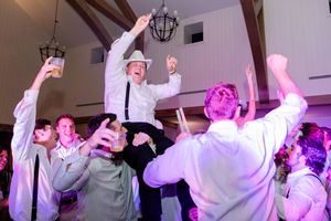 Wedding guests dance and celebrate on a crowded dance floor illuminated by purple and pink lighting at a reception.