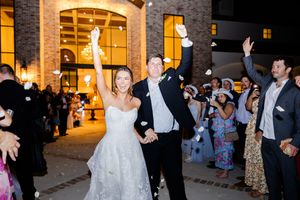 A wedding couple waves goodbye to guests during their evening reception exit at an illuminated stone venue.