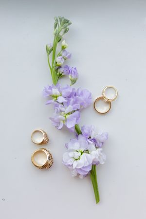 Gold wedding rings sit alongside delicate purple stock flowers against a light gray background in a minimalist arrangement.