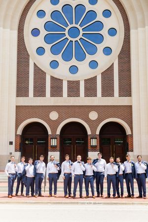 Wedding party lined up against ornate brick church backdrop with decorative blue circular window details above.