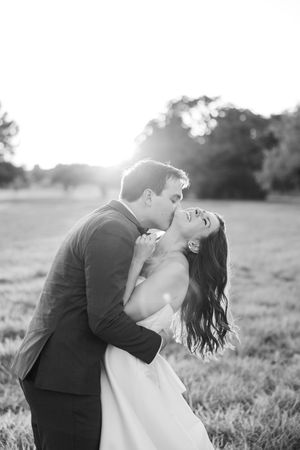 A couple in formal attire share romantic kisses in a sunlit field during a black and white photoshoot.