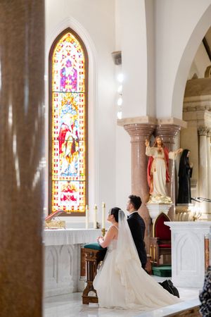 A couple kneels in prayer at a Catholic church altar with a beautiful stained glass window and religious statues in view.