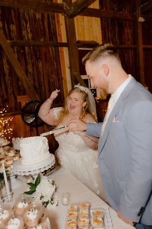 Bride and groom laughing together while cutting white wedding cake at rustic barn reception dessert table.