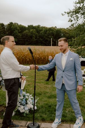 Groom in light blue suit shaking hands with officiant at outdoor wedding ceremony near a cornfield.