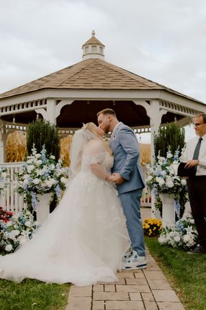 Bride and groom share first kiss at outdoor gazebo wedding ceremony surrounded by white and blue floral arrangements.