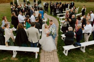 Aerial view of bride and groom kissing in aisle surrounded by standing wedding guests on white benches outdoors.