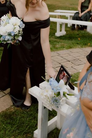 Bridesmaid in black off-shoulder dress placing memorial photo with blue hydrangeas on white ceremony bench.
