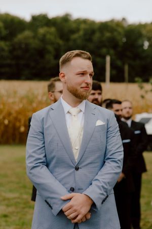 Emotional groom in light blue suit with teary eyes watching bride walk down the aisle outdoors.