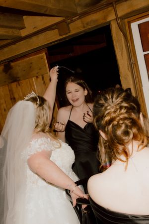 Bride in white gown dancing with guests in black dresses at a rustic wooden barn wedding reception.