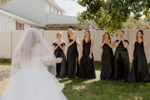 Bridesmaids in black dresses reacting with joy during bride first look in backyard.