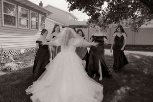 Black and white photo of bride twirling in wedding dress surrounded by bridesmaids outdoors.