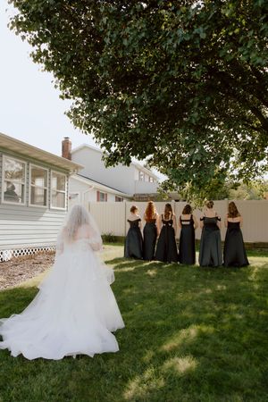 Bride in white gown with veil approaching bridesmaids in black dresses for first look.