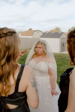Bride in lace wedding gown and veil makes a funny face while chatting with bridesmaids in black dresses outdoors.