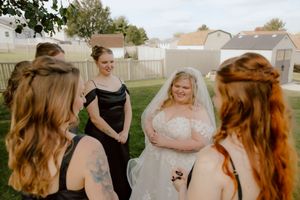 Bride in white lace gown laughs joyfully with bridesmaids in black dresses during outdoor wedding celebration.