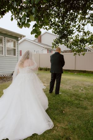 Bride in flowing white gown and veil approaches groom in black suit for first look in backyard garden.