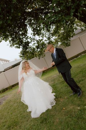 Bride in white ball gown dances playfully with groom under a tree during outdoor wedding first look.