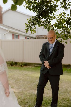 Emotional groom in black suit clasps hands and smiles tearfully during wedding first look reveal outdoors.