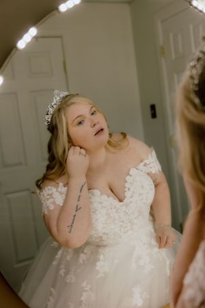 Bride in lace off-shoulder wedding gown and tiara adjusting earring while looking in a lighted mirror.
