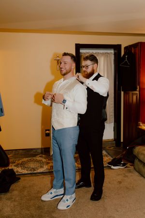 Groomsman helps groom adjust his white vest and blue wedding suit in a warmly lit indoor room.