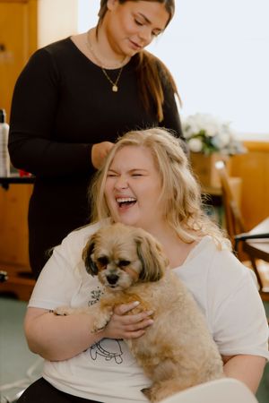Joyful bride laughs while holding her small fluffy dog as a stylist does her hair on wedding day.