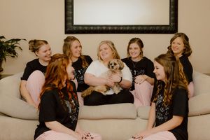 Bride surrounded by bridesmaids in black shirts on a couch, holding an adorable small dog during wedding preparations.
