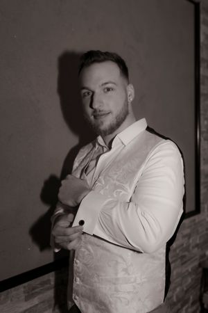 Dapper groomsman poses in a white patterned vest and dress shirt in a classic black and white photo.