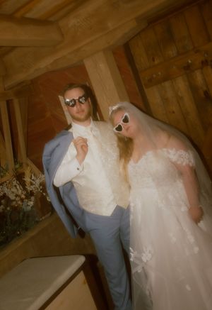 Bride and groom posing playfully in a rustic barn wearing stylish sunglasses, bride in lace gown with veil, groom in blue suit.