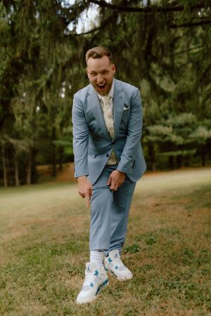 Laughing groom in light blue suit shows off fun white sneakers with blue accents on wedding day outdoors.