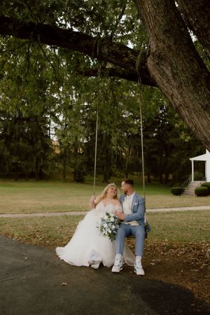 Bride and groom sit together on rustic rope swing beneath large oak tree during romantic outdoor wedding portraits.