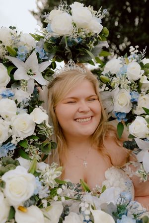 Smiling bride in tiara peeks through stunning arrangement of white roses and blue hydrangeas on wedding day.