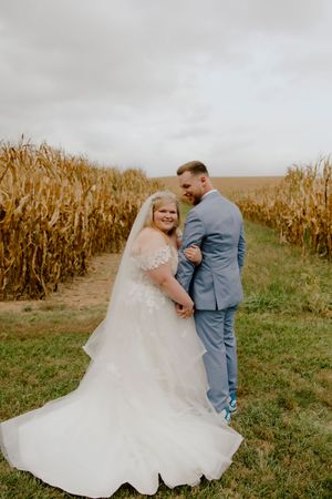 Bride and groom embracing in a cornfield pathway, bride wearing lace off-shoulder gown with veil, groom in light blue suit.