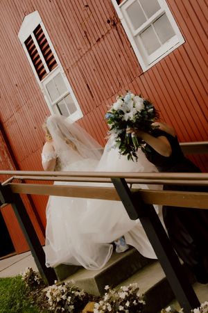Bride in white gown and veil carries bouquet while ascending stairs of a rustic red barn wedding venue.