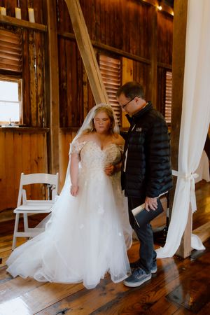 Bride in lace wedding gown receives guidance from officiant inside a warmly lit rustic barn wedding venue.