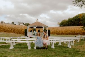 Two flower girls stand near a white gazebo surrounded by golden cornfields at an outdoor fall wedding ceremony.