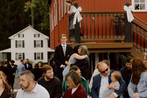 Wedding guests mingle outside a red barn venue while a couple embraces on the deck above.