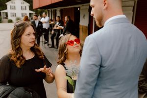 Wedding guests in black attire converse outdoors near rustic barn venue, one wearing bold red sunglasses.