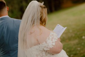 Bride in lace off-shoulder gown reads wedding vows from a card during an intimate outdoor ceremony.