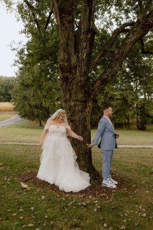 Bride in white lace gown and groom in blue suit share a first look moment beside a large tree.