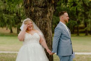 Emotional bride laughs and wipes tears during a touching first look moment with groom by a tree.