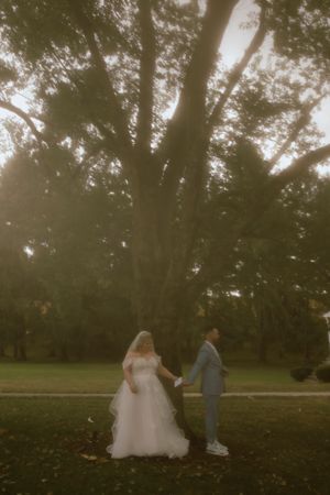 Bride and groom hold hands under a majestic tree in a dreamy, golden-lit outdoor wedding portrait.