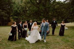 Bride and groom walk hand in hand with wedding party through green meadow surrounded by tall pine trees.